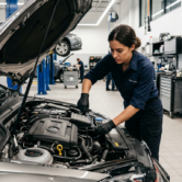 Website für Kfz-Werkstatt 1 Eine Kfz-Mechatronikerin arbeitet in einer modernen Autowerkstatt am geöffneten Motorraum eines Fahrzeugs. Sie trägt Arbeitskleidung und Handschuhe und prüft konzentriert Bauteile im Motorbereich. Im Hintergrund sind Hebebühnen, weitere Fahrzeuge, Werkzeugwagen, Regale mit Werkstattbedarf und ein weiterer Mitarbeiter zu sehen, wodurch die Szene professionelle Fahrzeugwartung, Reparatur und Service in einer gut ausgestatteten Kfz-Werkstatt vermittelt.