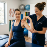 Website für Physiotherapeuten & Heilpraktiker 1 Eine Physiotherapeutin untersucht in einem hellen Behandlungsraum vorsichtig den Arm und die Schulter einer sitzenden Patientin. Im Hintergrund sind eine Behandlungsliege, Trainingsbälle, Faszienrollen, Übungsbänder und ein Anatomieposter zu sehen, wodurch die Szene eine professionelle physiotherapeutische Beratung oder manuelle Untersuchung vermittelt.