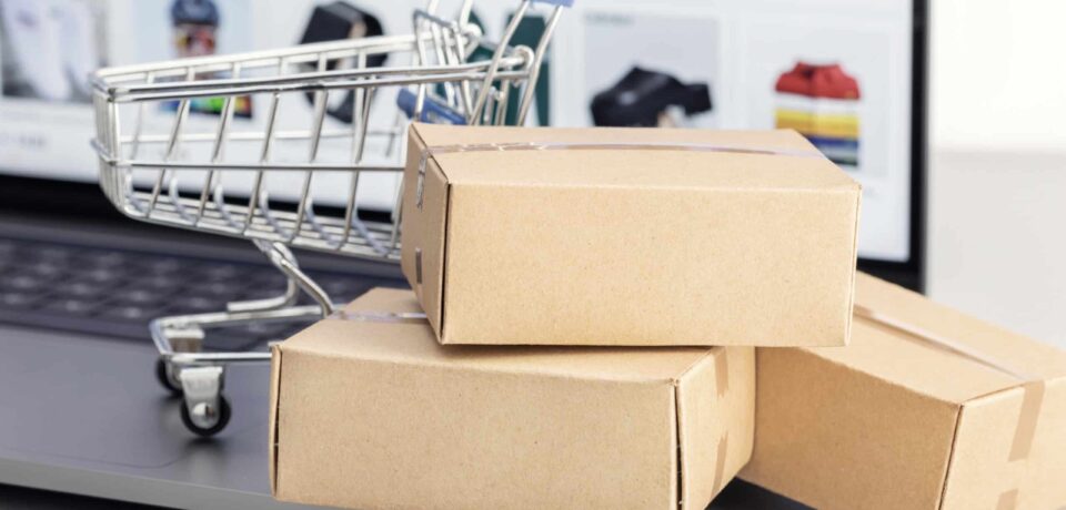 Mehrere Versandkartons liegen vor einem Laptop mit geöffnetem Onlineshop, daneben steht ein kleiner Einkaufswagen. Die Szene symbolisiert Onlinebestellungen, Warenkorb, Versand und E-Commerce-Prozesse. Das Bild passt zu Themen wie Onlineshop ohne Verkäufe, Kaufabbrüche, Warenkorb-Optimierung, Versandvertrauen und bessere Shop-Conversion.