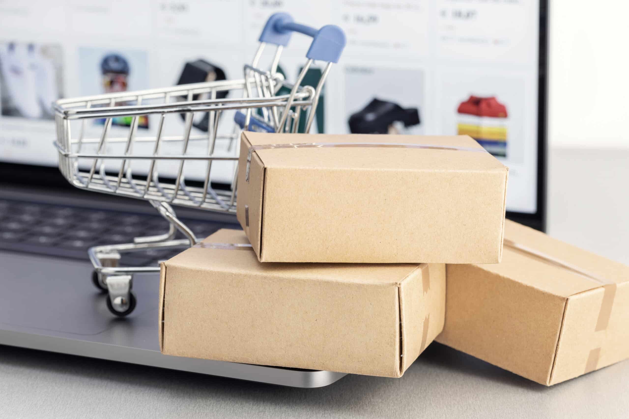 Mehrere Versandkartons liegen vor einem Laptop mit geöffnetem Onlineshop, daneben steht ein kleiner Einkaufswagen. Die Szene symbolisiert Onlinebestellungen, Warenkorb, Versand und E-Commerce-Prozesse. Das Bild passt zu Themen wie Onlineshop ohne Verkäufe, Kaufabbrüche, Warenkorb-Optimierung, Versandvertrauen und bessere Shop-Conversion.