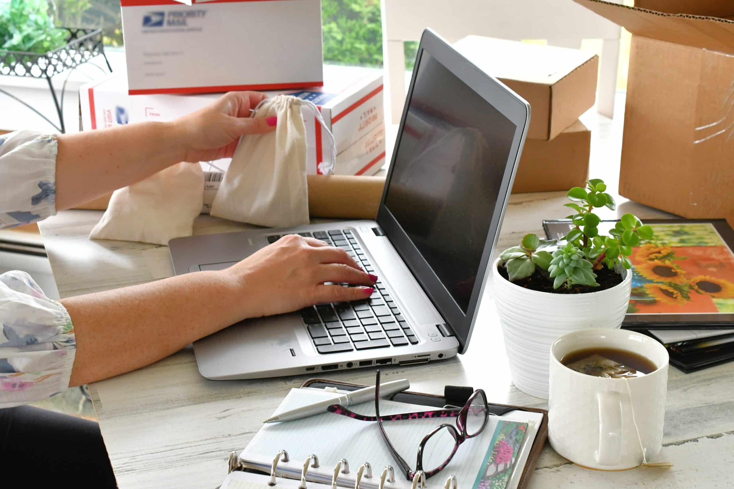 Eine Person arbeitet am Laptop und bereitet kleine Stoffbeutel für den Versand vor. Im Hintergrund stehen Kartons und Versandmaterial, daneben liegen Notizbuch, Stift, Brille, Pflanze und eine Tasse. Das Bild passt zu Themen wie Onlineshop betreiben, Bestellungen bearbeiten, Versand vorbereiten, Produktverpackung, kleine Shops und E-Commerce-Alltag.