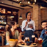 Eine Servicekraft nimmt in einem gemütlichen Restaurant mit Tablet Bestellungen von Gästen auf. Das Bild steht für moderne Gastronomie, digitale Bestellaufnahme, professionellen Service, Restaurantbetrieb, Reservierungen und digitale Abläufe in Restaurants.