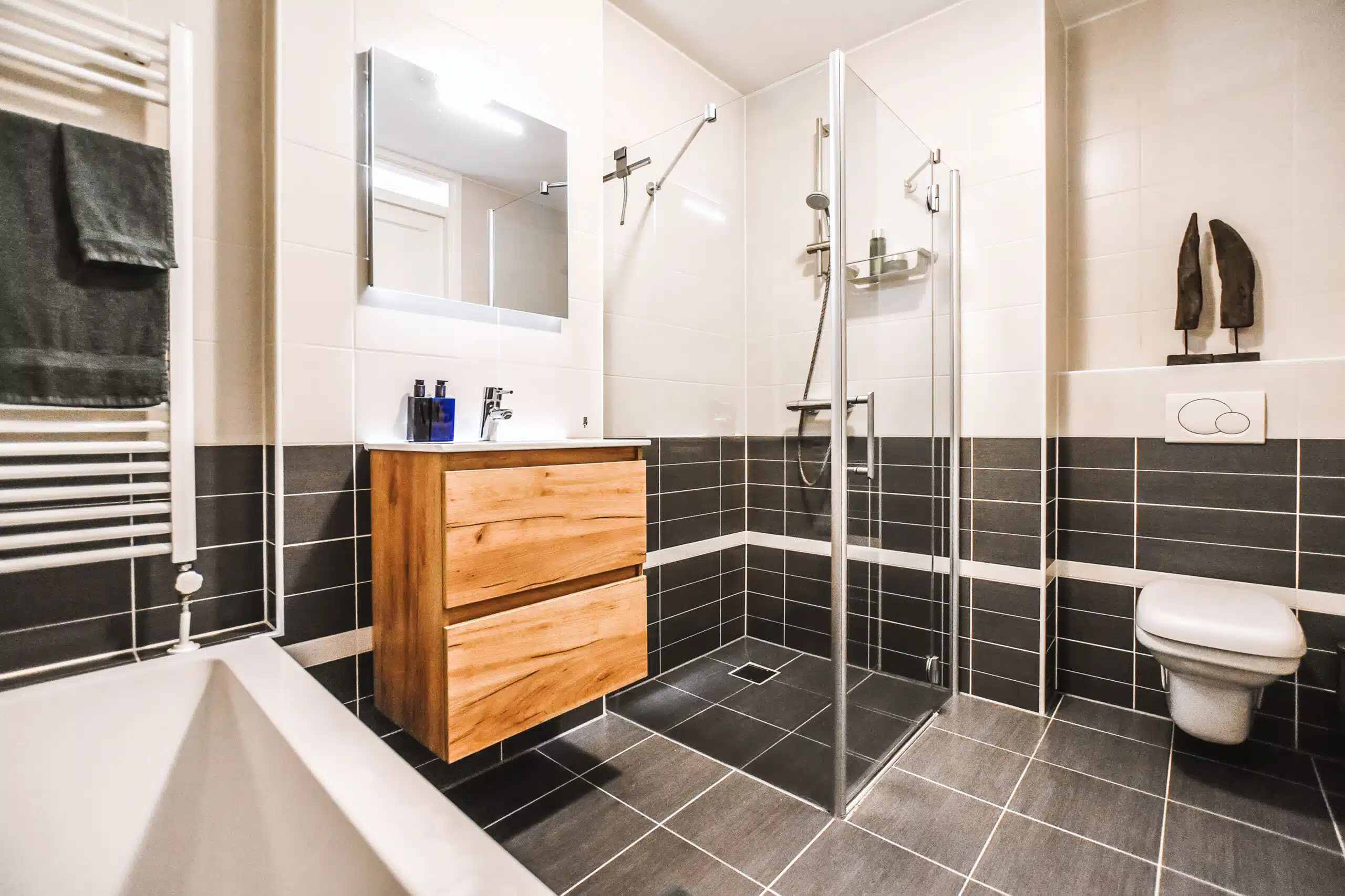 Modern saniertes Badezimmer mit schwarz-weißen Wand- und Bodenfliesen, Glasdusche, Badewanne, Holzwaschtisch, Spiegel und klarer zeitgemäßer Gestaltung.