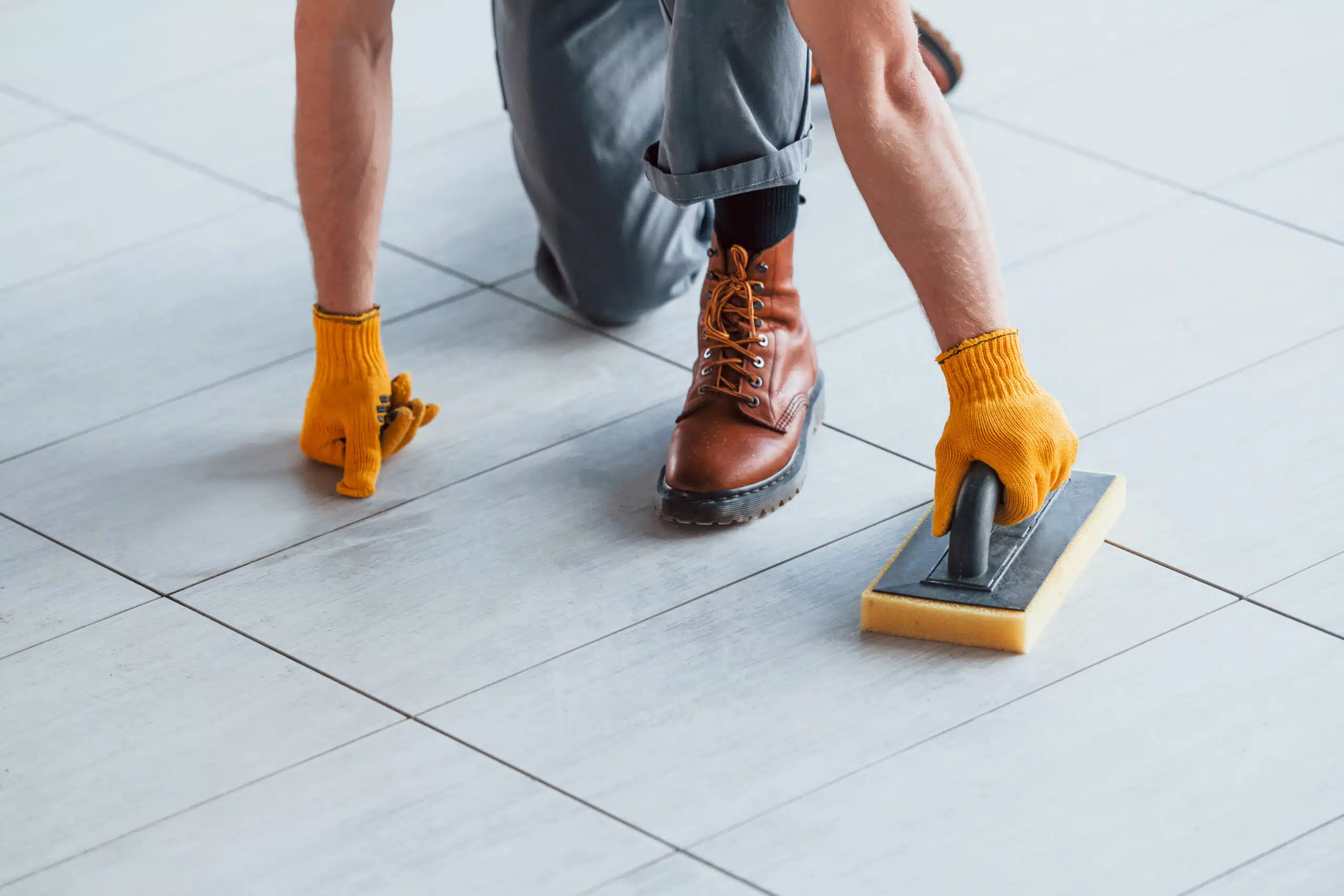 Handwerker reinigt oder verfugt helle Bodenfliesen mit einem Schwammbrett und trägt dabei gelbe Arbeitshandschuhe, während er kniend auf dem frisch verlegten Fliesenboden arbeitet.