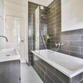 Delightful bathroom with a cozy bathtub Helles modernes Badezimmer mit grauen Wandfliesen, weißer Badewanne, Glas-Duschabtrennung, Waschbecken, großem Spiegel und sauber verarbeiteten Fliesenflächen.