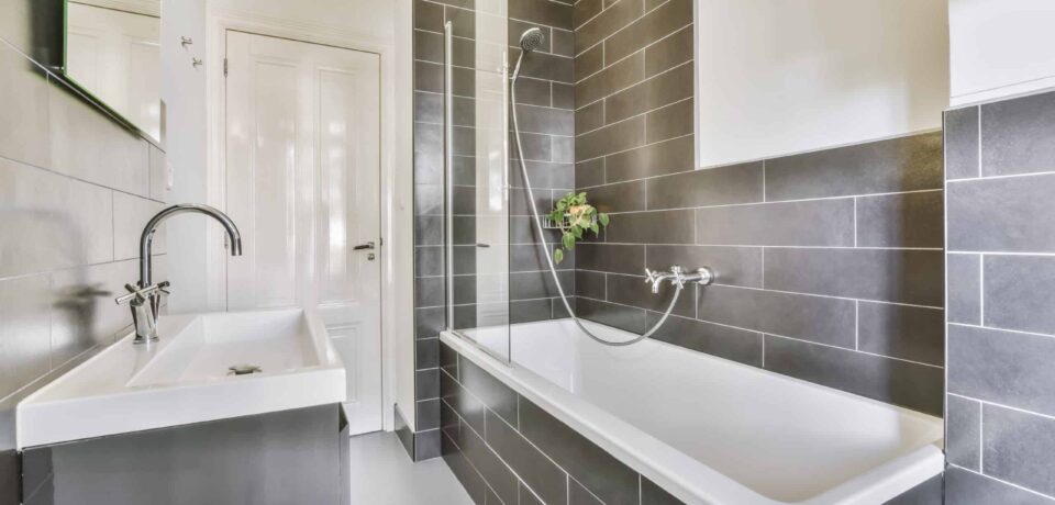 Delightful bathroom with a cozy bathtub Helles modernes Badezimmer mit grauen Wandfliesen, weißer Badewanne, Glas-Duschabtrennung, Waschbecken, großem Spiegel und sauber verarbeiteten Fliesenflächen.