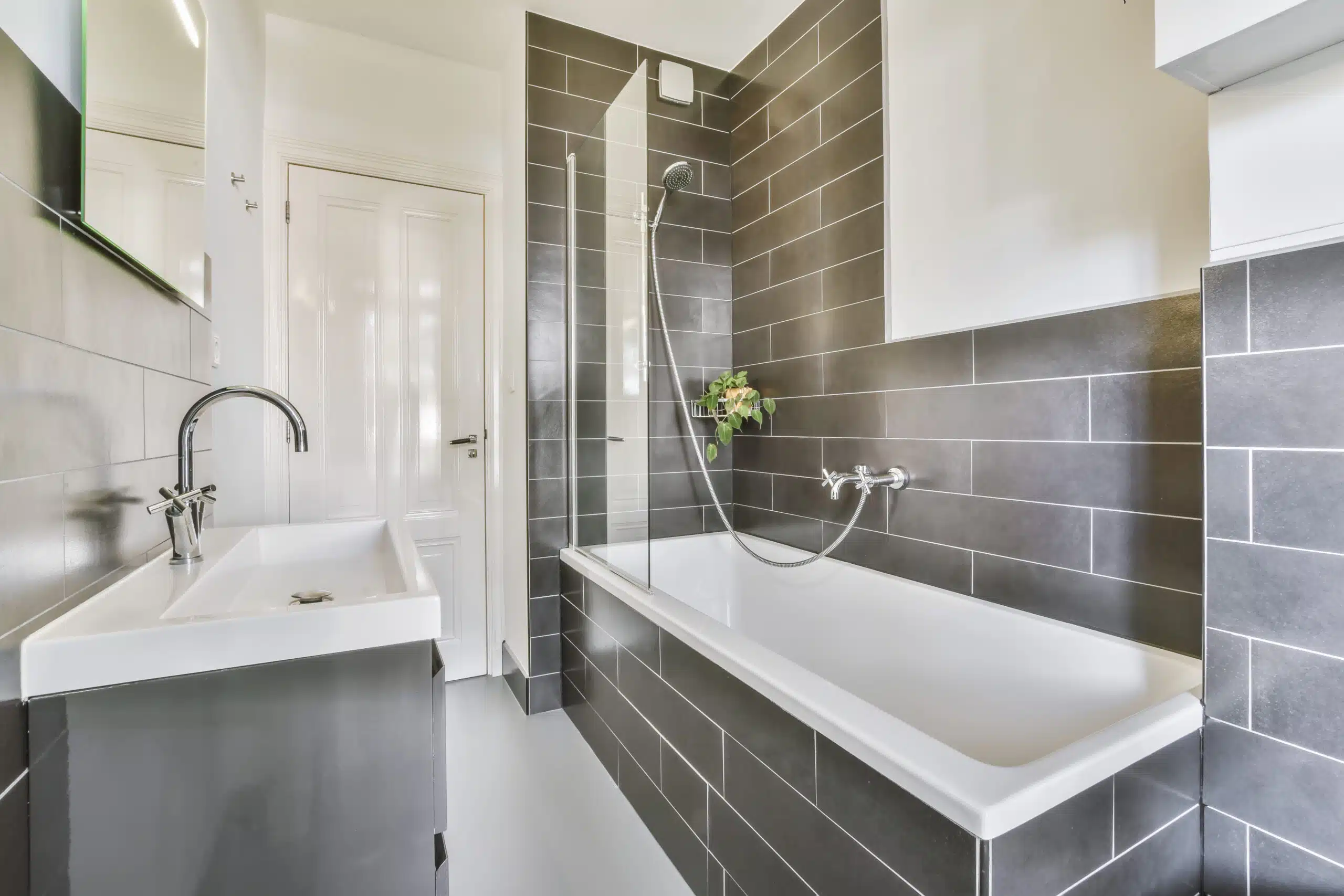 Helles modernes Badezimmer mit grauen Wandfliesen, weißer Badewanne, Glas-Duschabtrennung, Waschbecken, großem Spiegel und sauber verarbeiteten Fliesenflächen.