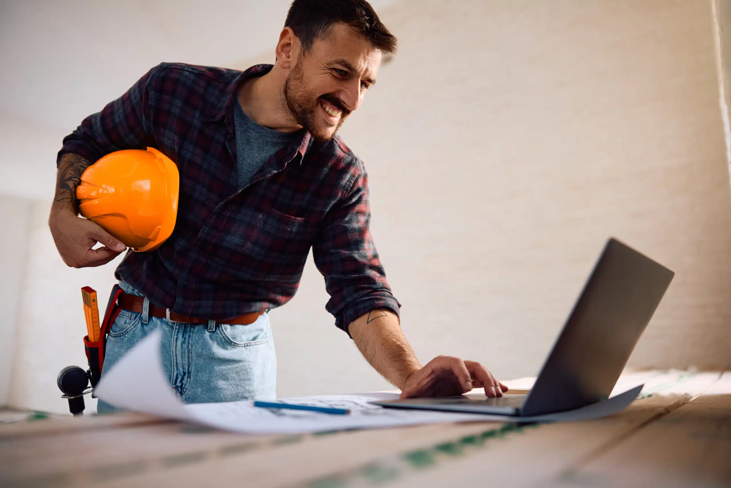 Fliesenleger auf einer Baustelle prüft mit einem Laptop digitale Baupläne und Projektunterlagen, während er einen orangefarbenen Schutzhelm unter dem Arm hält.